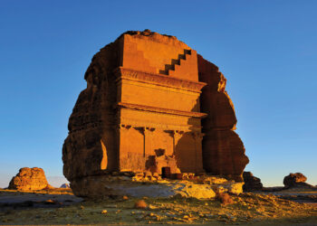 Qasr Al Farid, a Nabatean Tomb in Hegra, Saudi Arabia. Photo shows the tomb, known as the lonely castle, standing apart from the other structures against a blue background