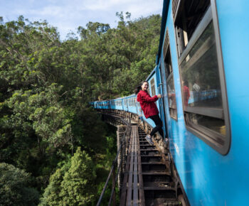 Asian girl with red jacked hanging from blue train during famous Ella-Kandy train ride